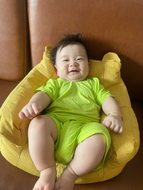 Bé Tú Tài cười trên gối vàng trong bộ đồ xanh